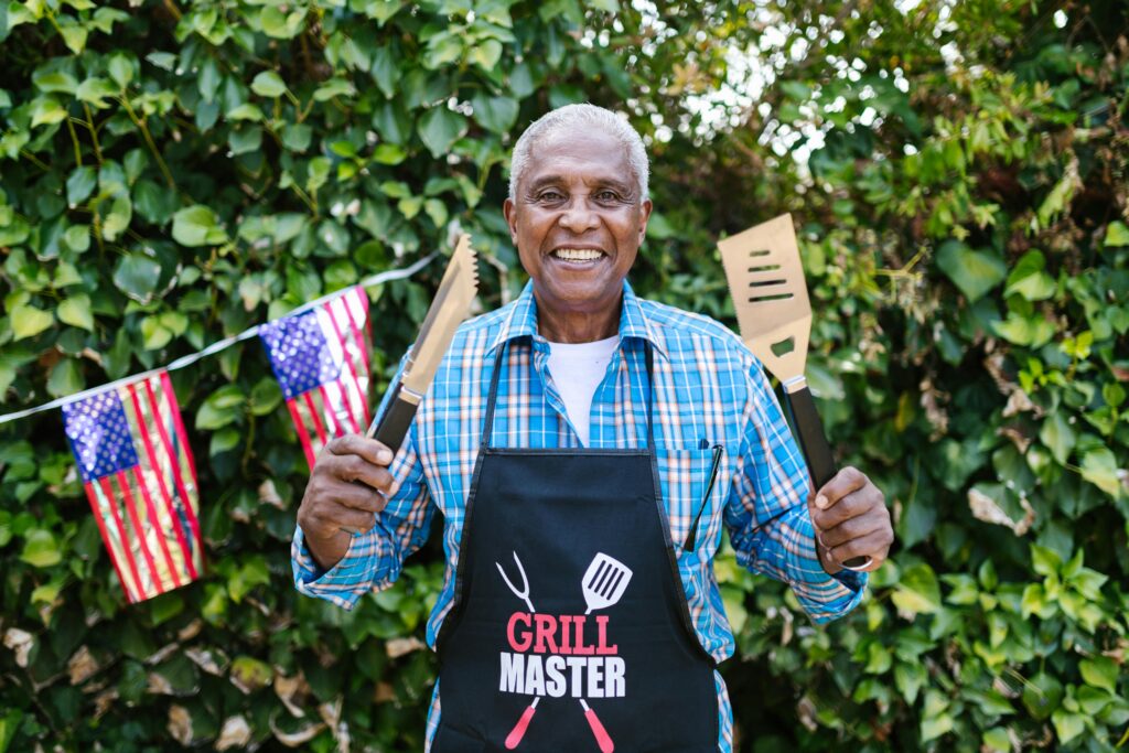 Black man holding up a spatula and tongs ready to grill with a grill master apron on